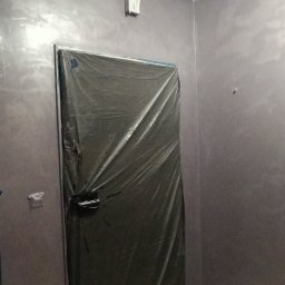 Dat-fix Radosław Data - Pokój w trakcie prac wykończeniowych, ściany pomalowane na szaro z efektem połysku, zabezpieczone folią drzwi, widoczna taśma malarska na suficie i przy podłodze, małe wiaderko z pędzlami przy...