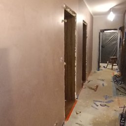 Dat-fix Radosław Data - Korytarz w trakcie prac wykończeniowych, ściany pokryte gładzią w odcieniach beżu, zabezpieczone framugi drzwi, podłoga z resztkami taśmy malarskiej i zabrudzeniami po remoncie.