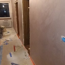 Dat-fix Radosław Data - Korytarz w trakcie prac wykończeniowych, ściany pokryte gładzią w odcieniach brązu, zabezpieczona podłoga z widocznymi narzędziami i poziomnicą.