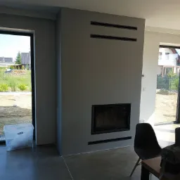 Szara ściana z wbudowanym kominkiem i czarnymi kratkami wentylacyjnymi, widok na zewnątrz przez okno, surowy beton na podłodze, krzesło i stół w pokoju, w tle dom w budowie.