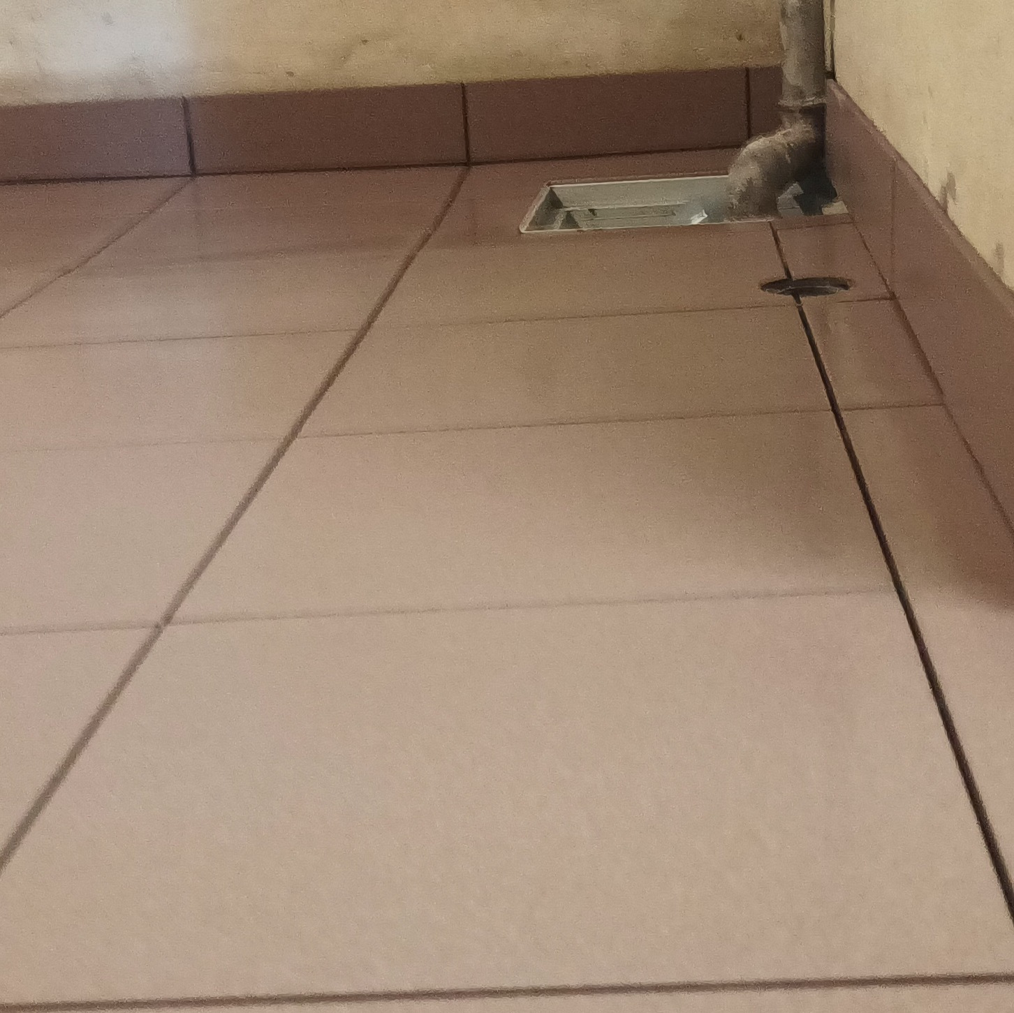 Beżowe płytki podłogowe z widocznym odpływem liniowym i rurą kanalizacyjną przy ścianie, ukazujące detale wykończenia łazienki.