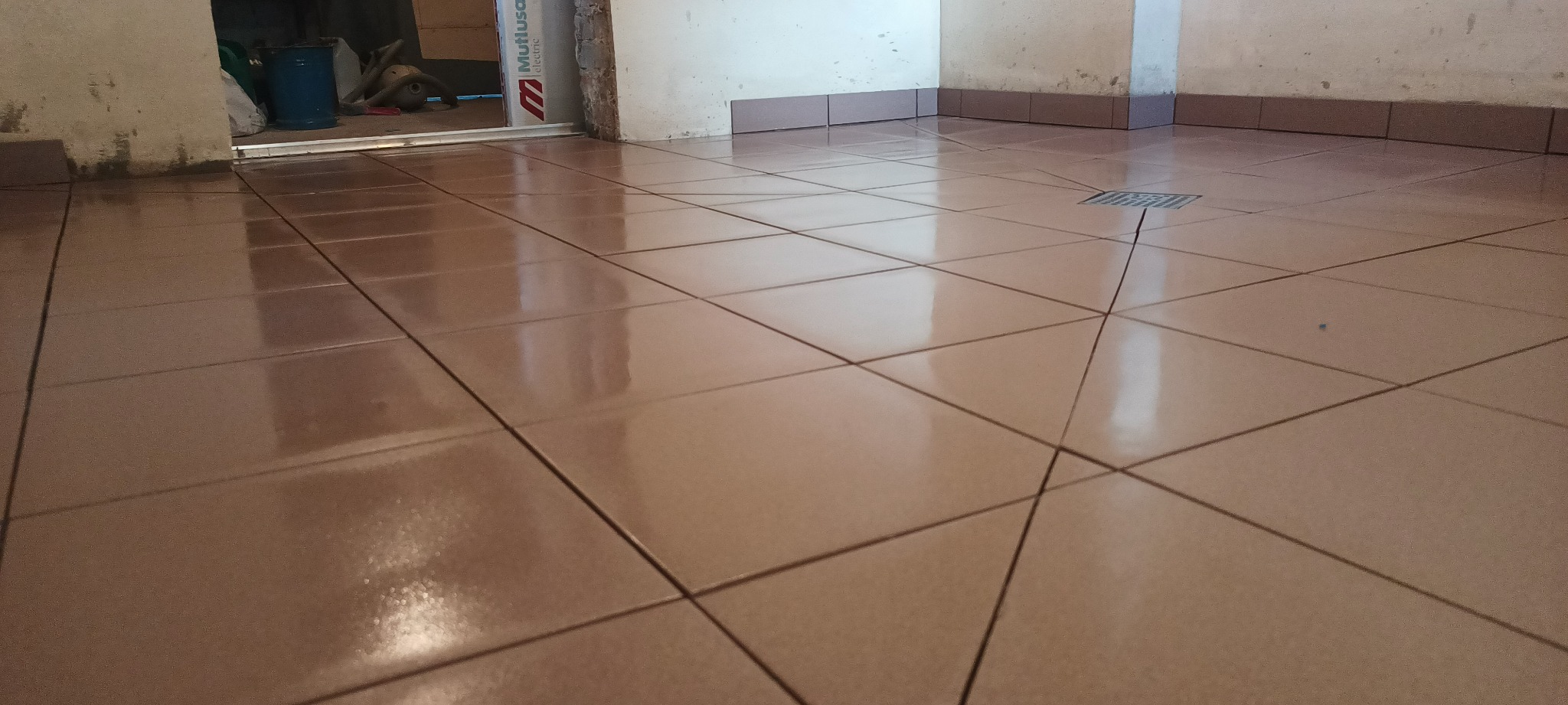 Świeżo położona, połyskująca, beżowa podłoga z płytek ceramicznych z odpływem liniowym w rogu, widoczne narzędzia i materiały budowlane w tle.