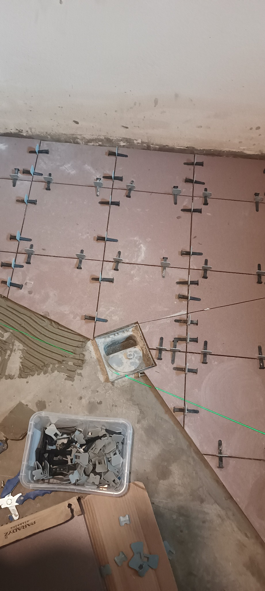 Układanie brązowych płytek podłogowych z użyciem systemu poziomowania, widoczny odpływ podłogowy i zielony laser krzyżowy.