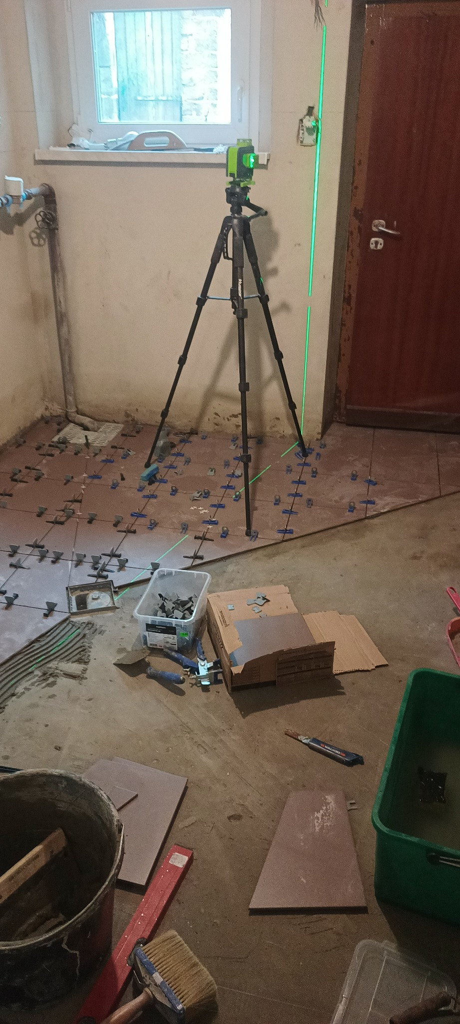 Układanie brązowych płytek podłogowych z użyciem lasera krzyżowego i plastikowych krzyżyków dystansowych, widoczne narzędzia i akcesoria glazurnicze, pomieszczenie w trakcie remontu.