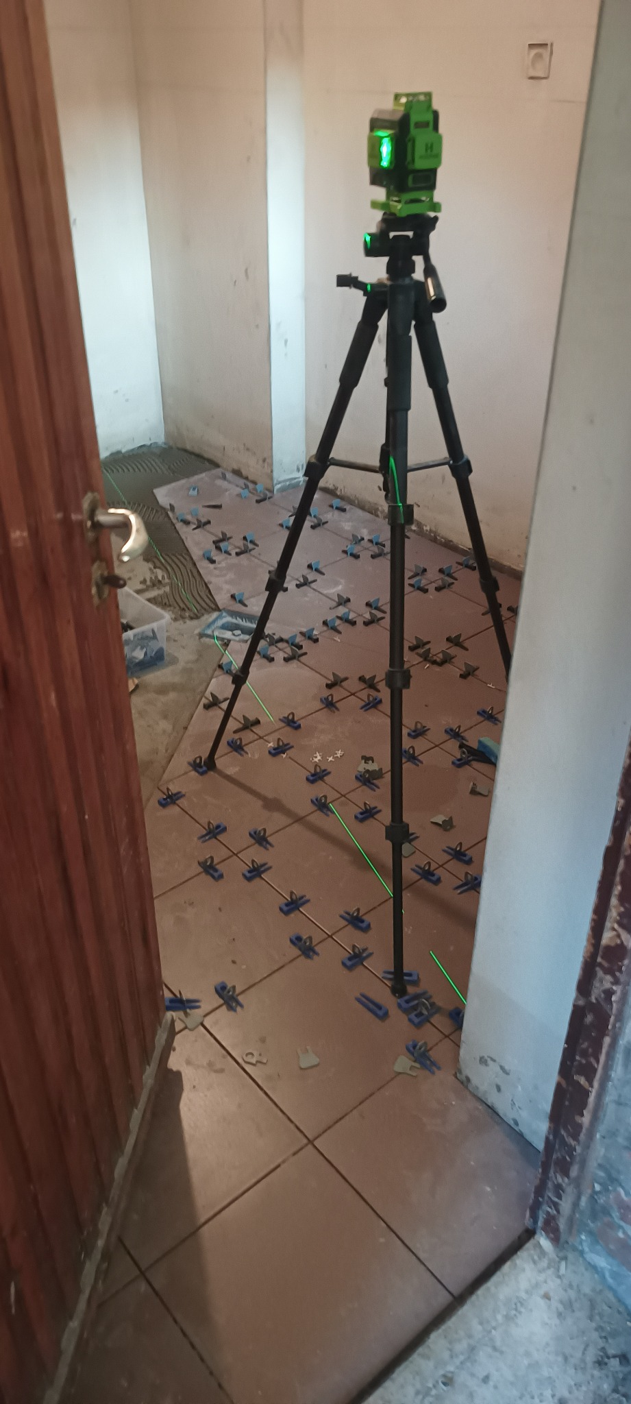 Układanie płytek podłogowych z użyciem poziomicy laserowej i systemu poziomowania płytek, widoczne krzyżyki i kliny, pomieszczenie w trakcie remontu.