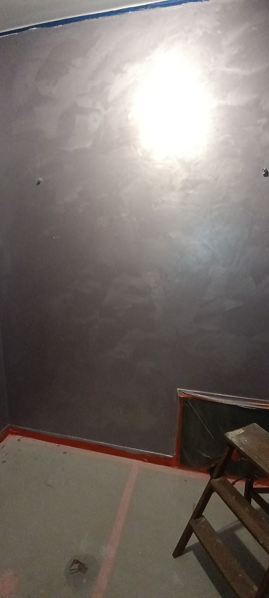 Ściana pokryta dekoracyjną, szarą powłoką z efektem chmur, zabezpieczona taśmą malarską przy suficie i podłodze, z widoczną drabinką malarską i folią ochronną.