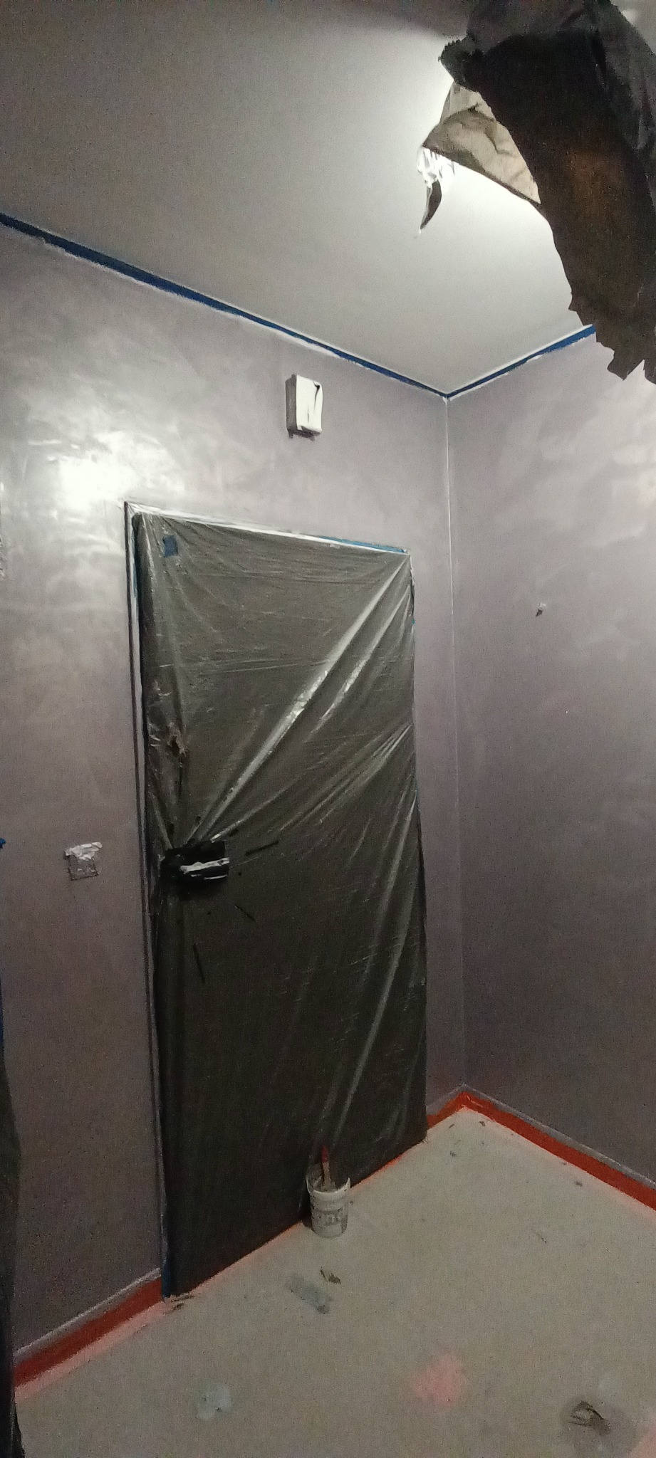 Pokój w trakcie prac wykończeniowych, ściany pomalowane na szaro z efektem połysku, zabezpieczone folią drzwi, widoczna taśma malarska na suficie i przy podłodze, małe wiaderko z pędzlami przy...