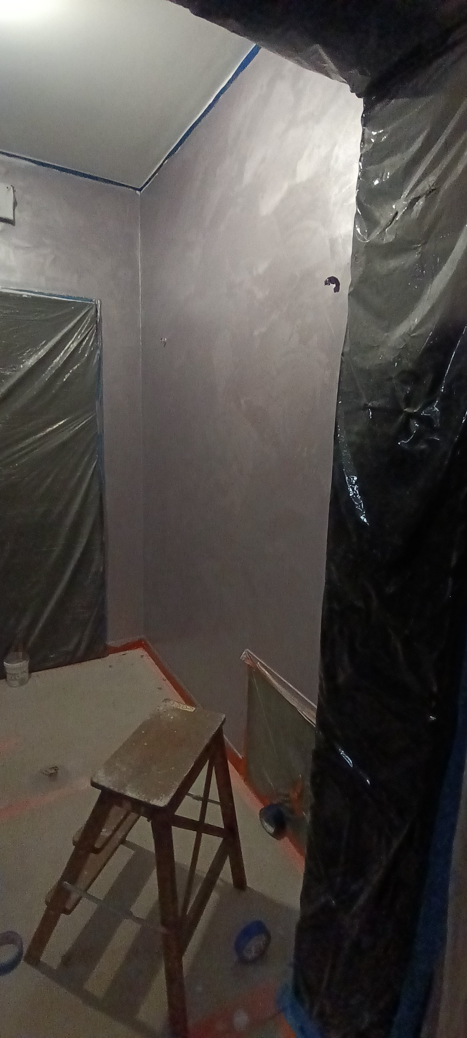 Świeżo pomalowane ściany w odcieniach szarości z delikatnym efektem dekoracyjnym, zabezpieczone folią meble i schodki malarskie w pomieszczeniu w trakcie prac wykończeniowych.