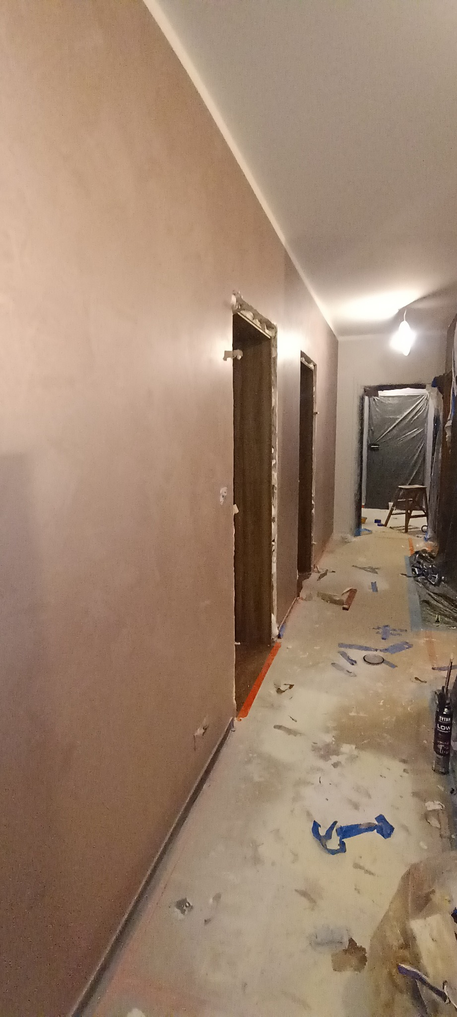 Korytarz w trakcie prac wykończeniowych, ściany pokryte gładzią w odcieniach beżu, zabezpieczone framugi drzwi, podłoga z resztkami taśmy malarskiej i zabrudzeniami po remoncie.