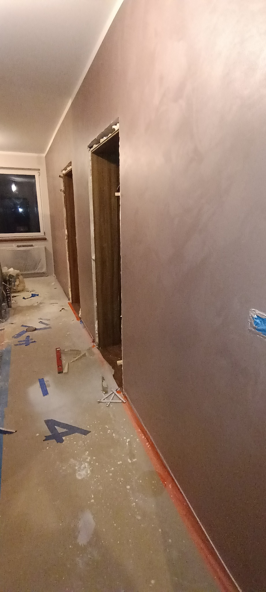 Korytarz w trakcie prac wykończeniowych, ściany pokryte gładzią w odcieniach brązu, zabezpieczona podłoga z widocznymi narzędziami i poziomnicą.
