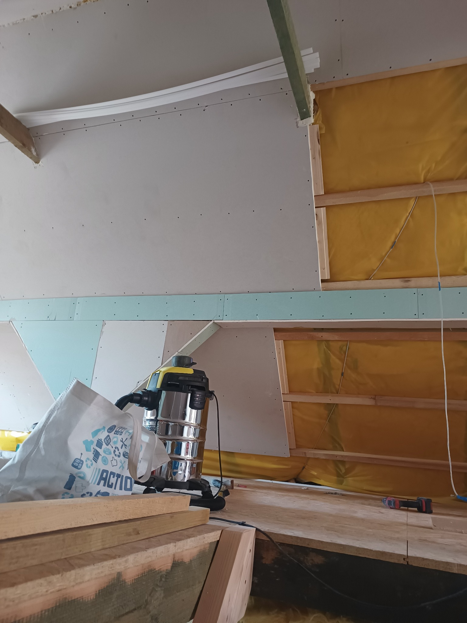 Montaż płyty karton gips na poddaszu z widoczną izolacją, metalowym odkurzaczem przemysłowym i workiem firmy Action.