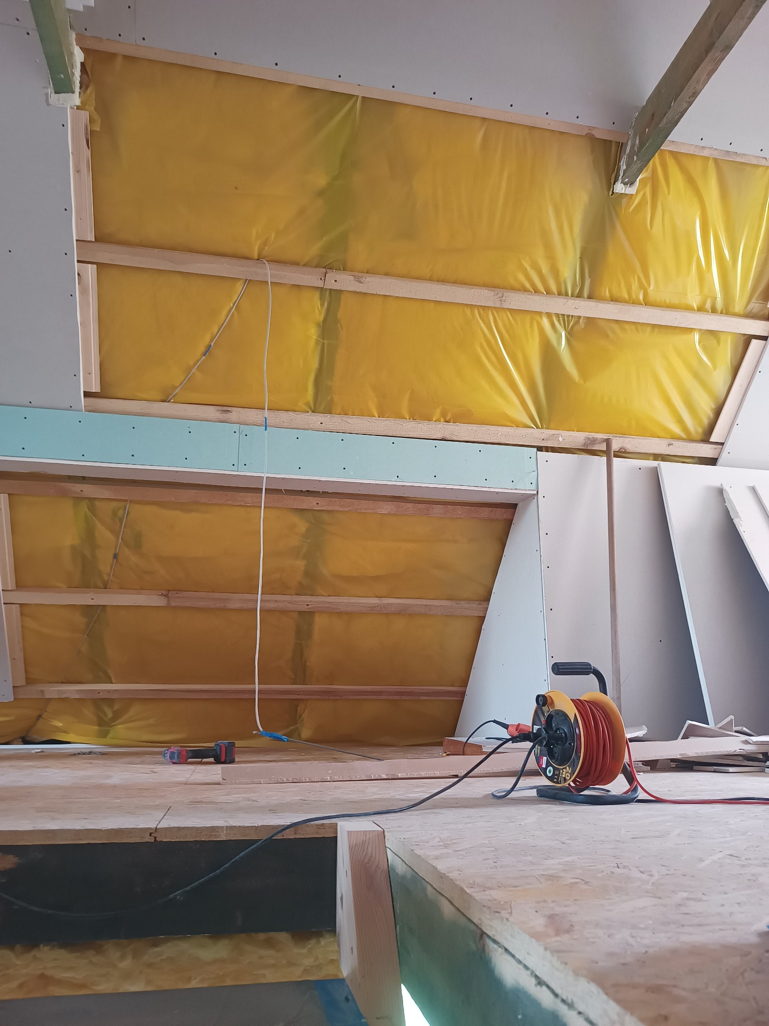 Poddasze w trakcie adaptacji: widoczne ocieplenie folią, konstrukcja drewniana, częściowo zamontowane płyty gipsowo-kartonowe, przedłużacz zwinięty na bębnie, narzędzia na podłodze.