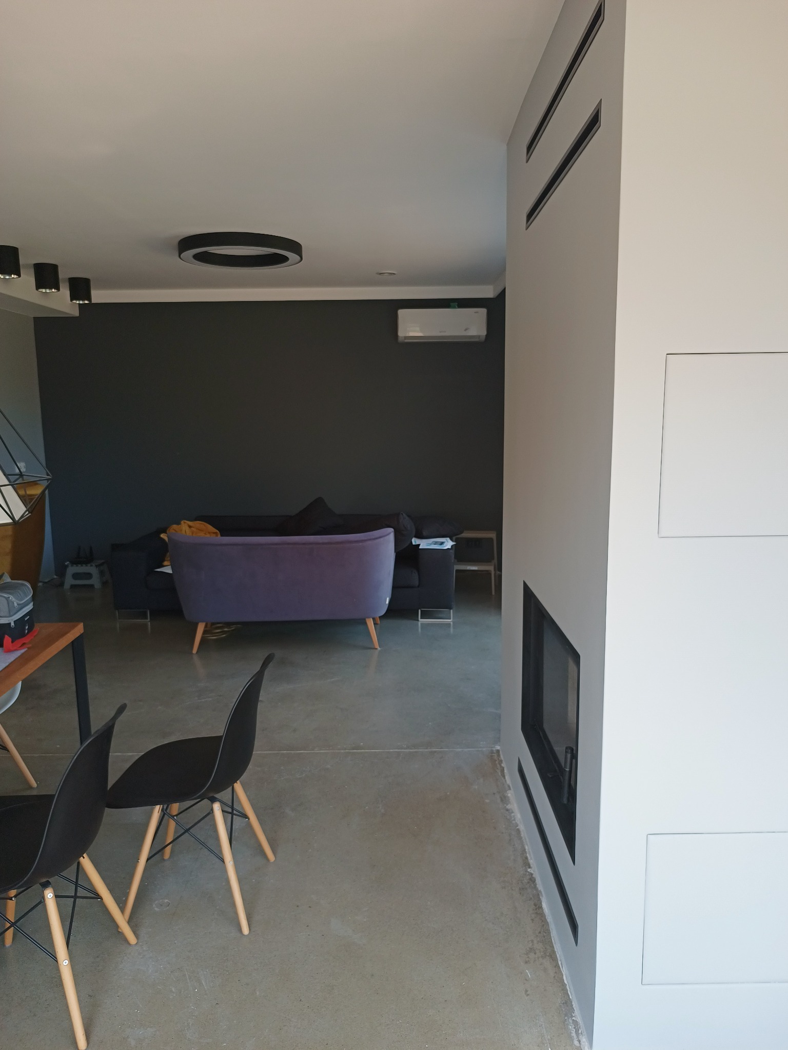 Nowoczesne wnętrze salonu z szarą ścianą, minimalistycznym kominkiem, betonową podłogą i designerskimi krzesłami.