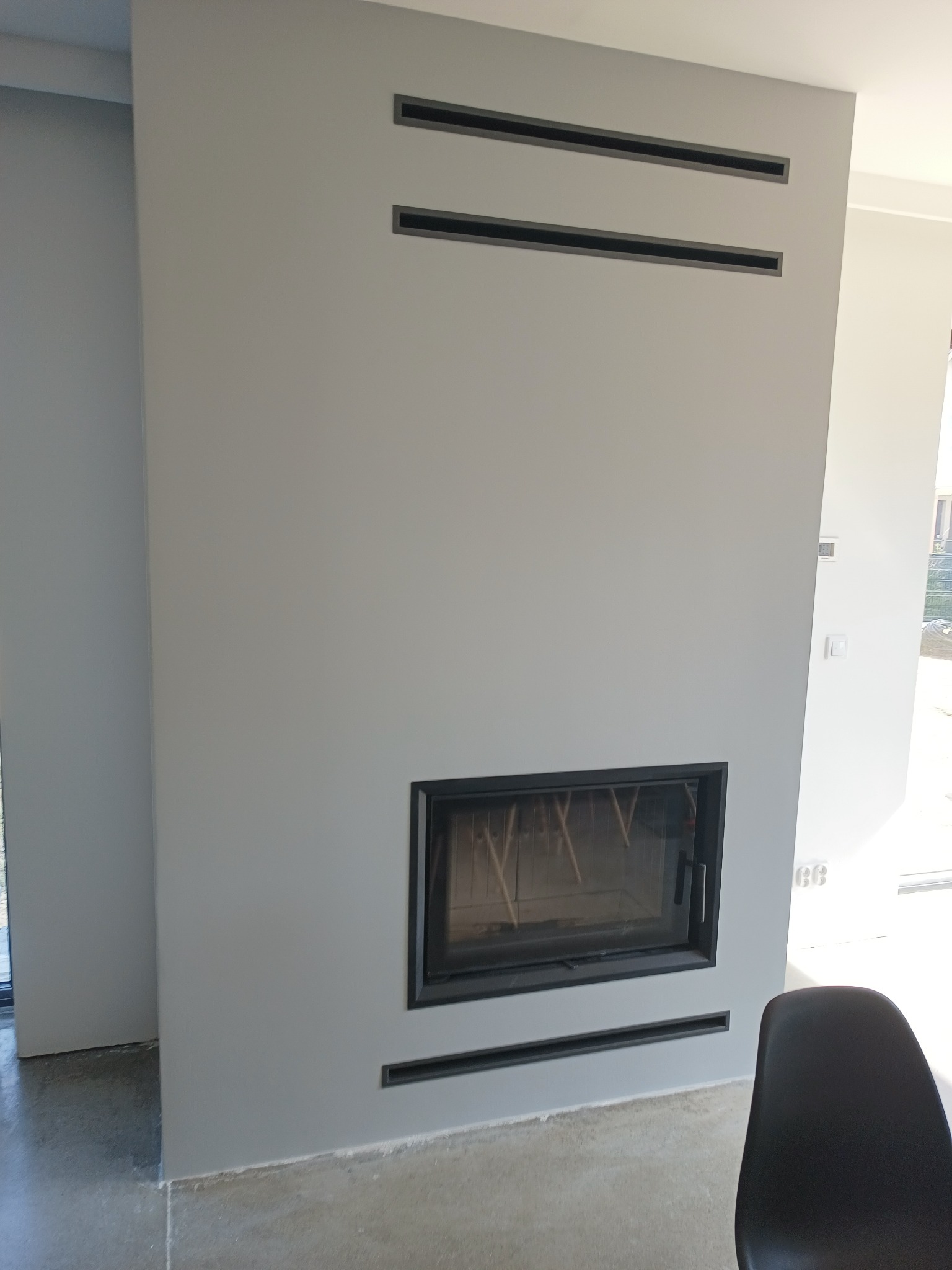 Minimalistyczny kominek w odcieniach szarości z czarną obudową i poziomymi szczelinami wentylacyjnymi, częściowo widoczne krzesło na pierwszym planie, surowa podłoga.