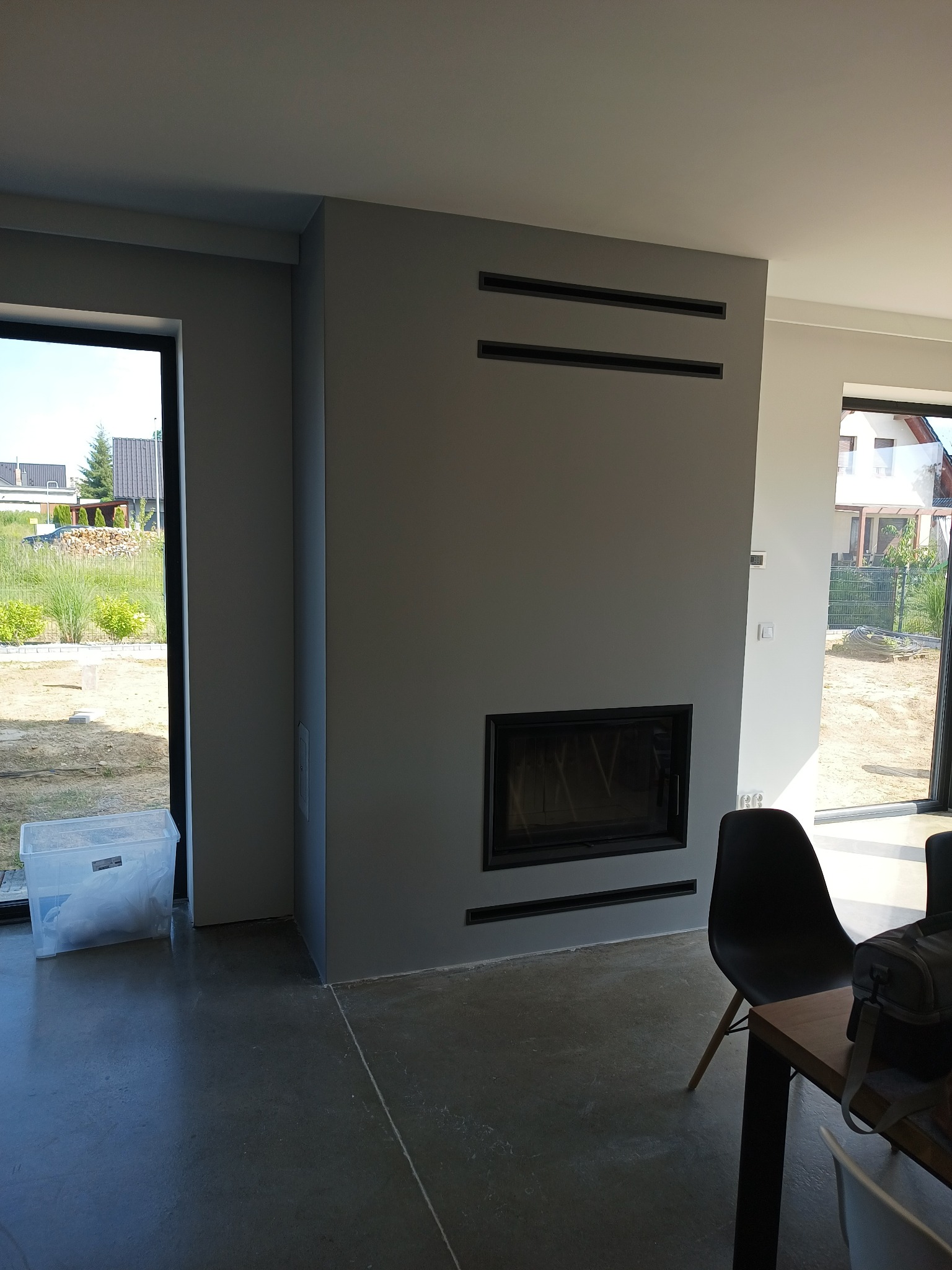 Szara ściana z wbudowanym kominkiem i czarnymi kratkami wentylacyjnymi, widok na zewnątrz przez okno, surowy beton na podłodze, krzesło i stół w pokoju, w tle dom w budowie.