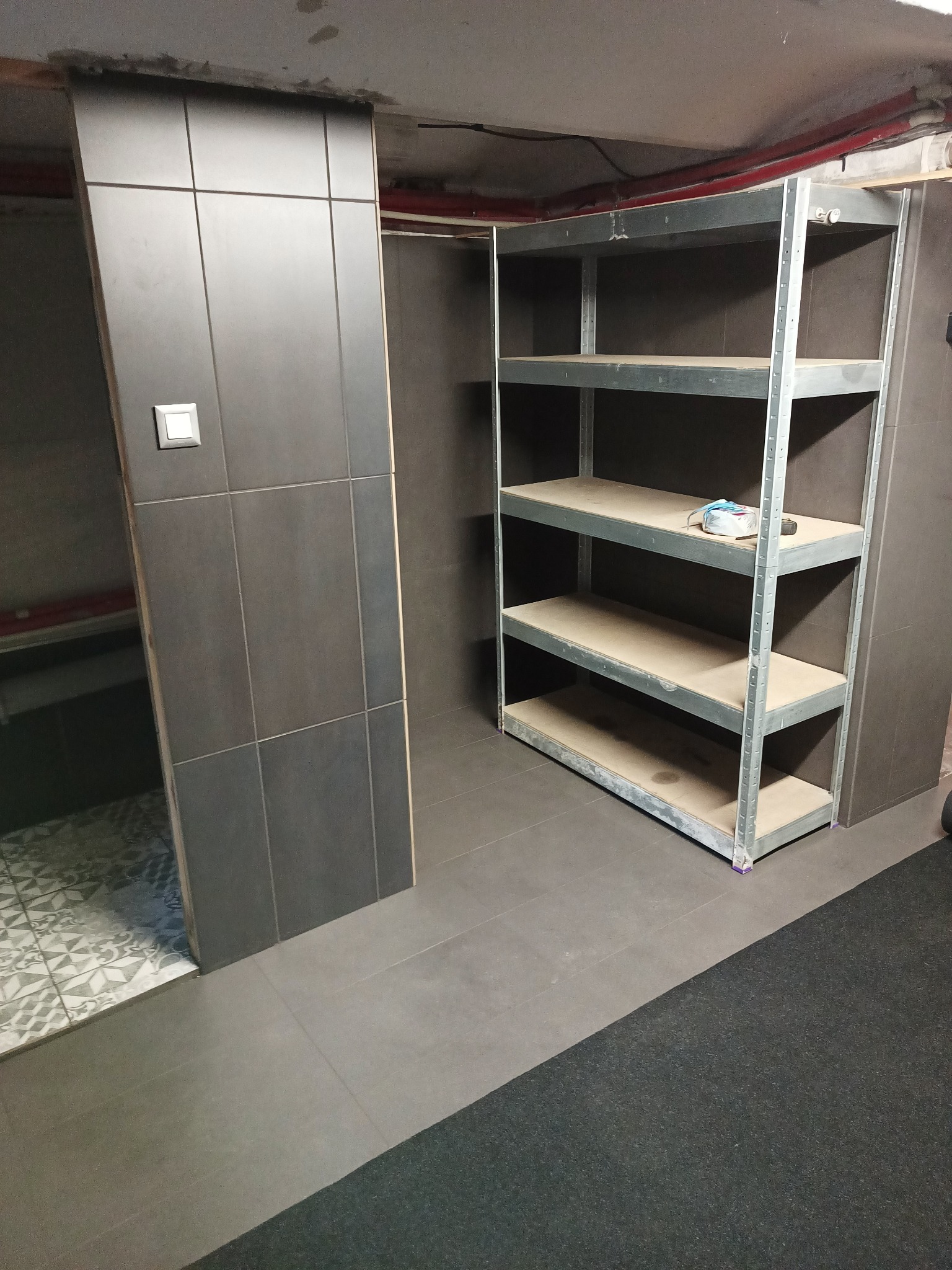 Wykończone wnętrze piwnicy z ciemnymi płytkami na ścianach i podłodze, metalowy regał z drewnianymi półkami stoi przy ścianie, a w tle widoczny fragment pomieszczenia z ozdobną podłogą.