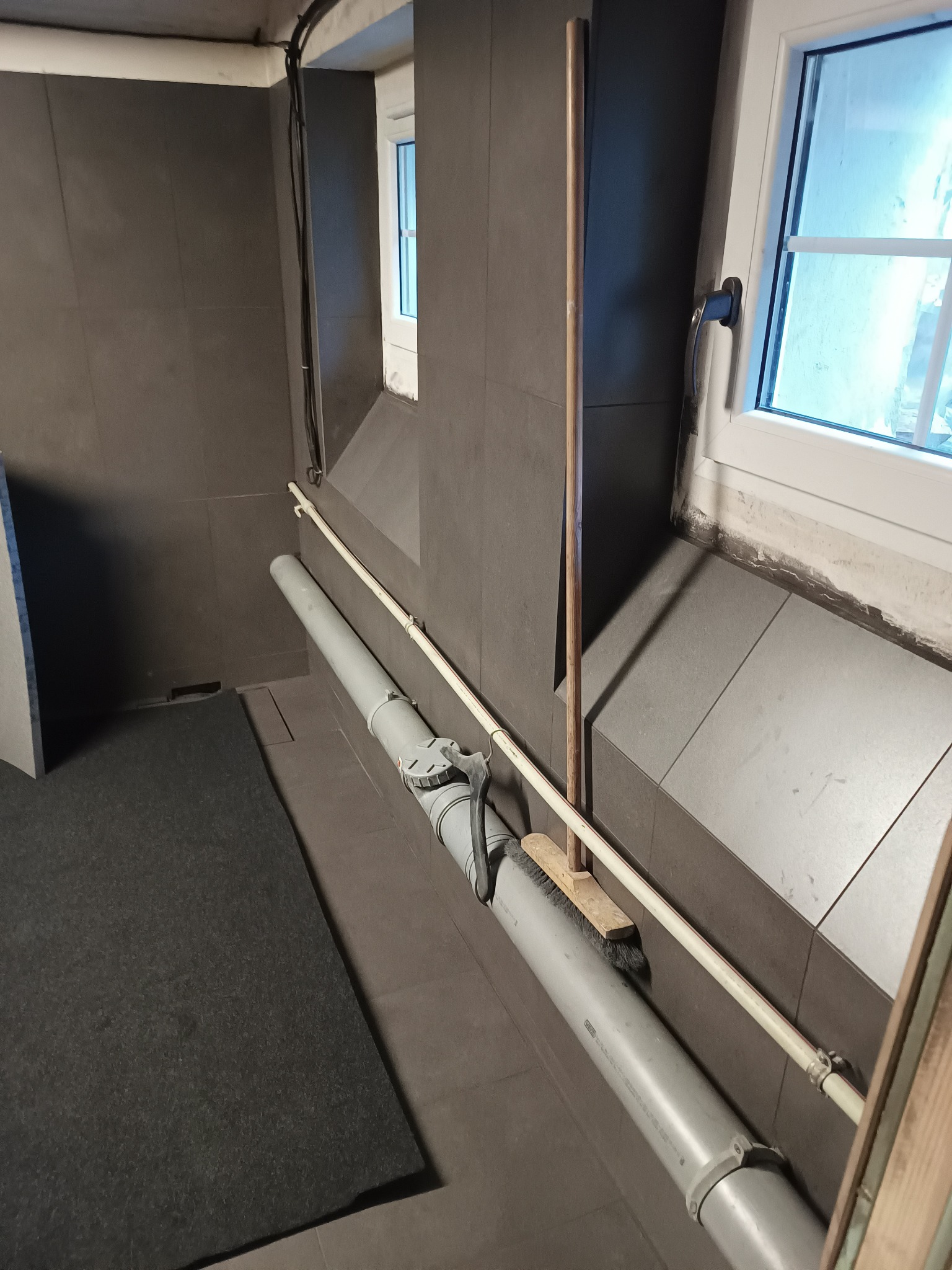 Szare płytki ścienne i podłogowe w piwnicy, rura kanalizacyjna z zamontowaną szczotką i drewnianym kijem opartym o ścianę, okno z widocznymi śladami prac wykończeniowych.