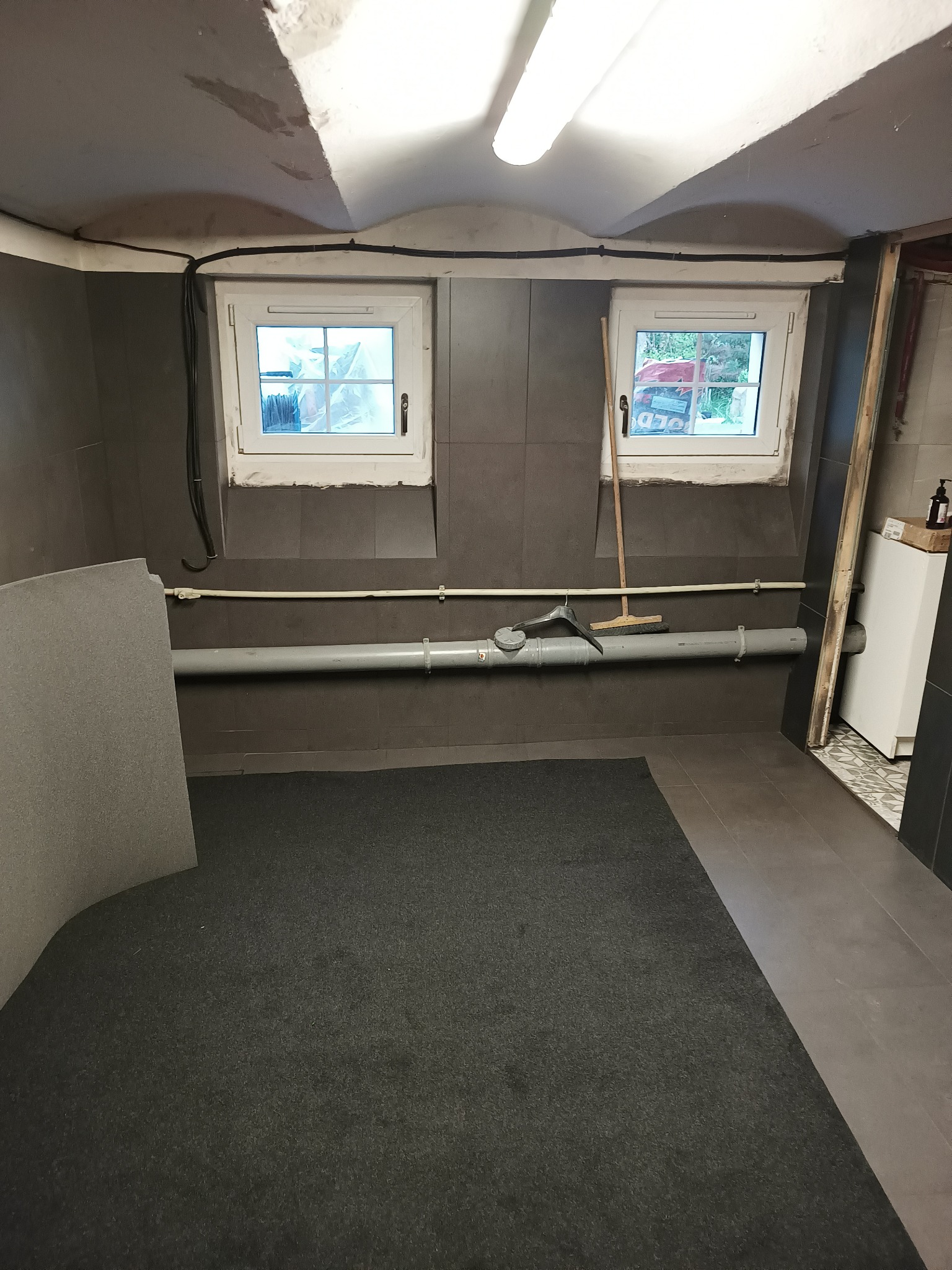 Piwnica z szarą podłogą, płytami kartonowo-gipsowymi opartymi o ścianę, rurami pod sufitem, oknami i lampą, w trakcie remontu.