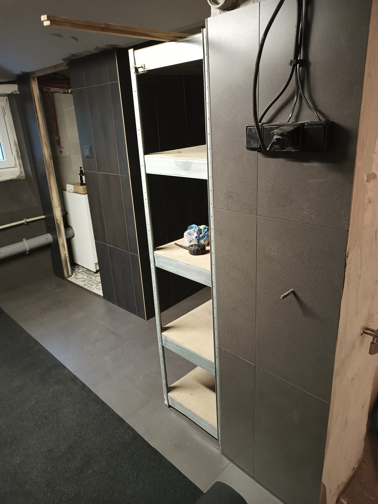 Półki regałowe w pomieszczeniu po remoncie w Mikołowie, widoczne szare płytki na ścianach, fragment pralki i instalacji elektrycznej.