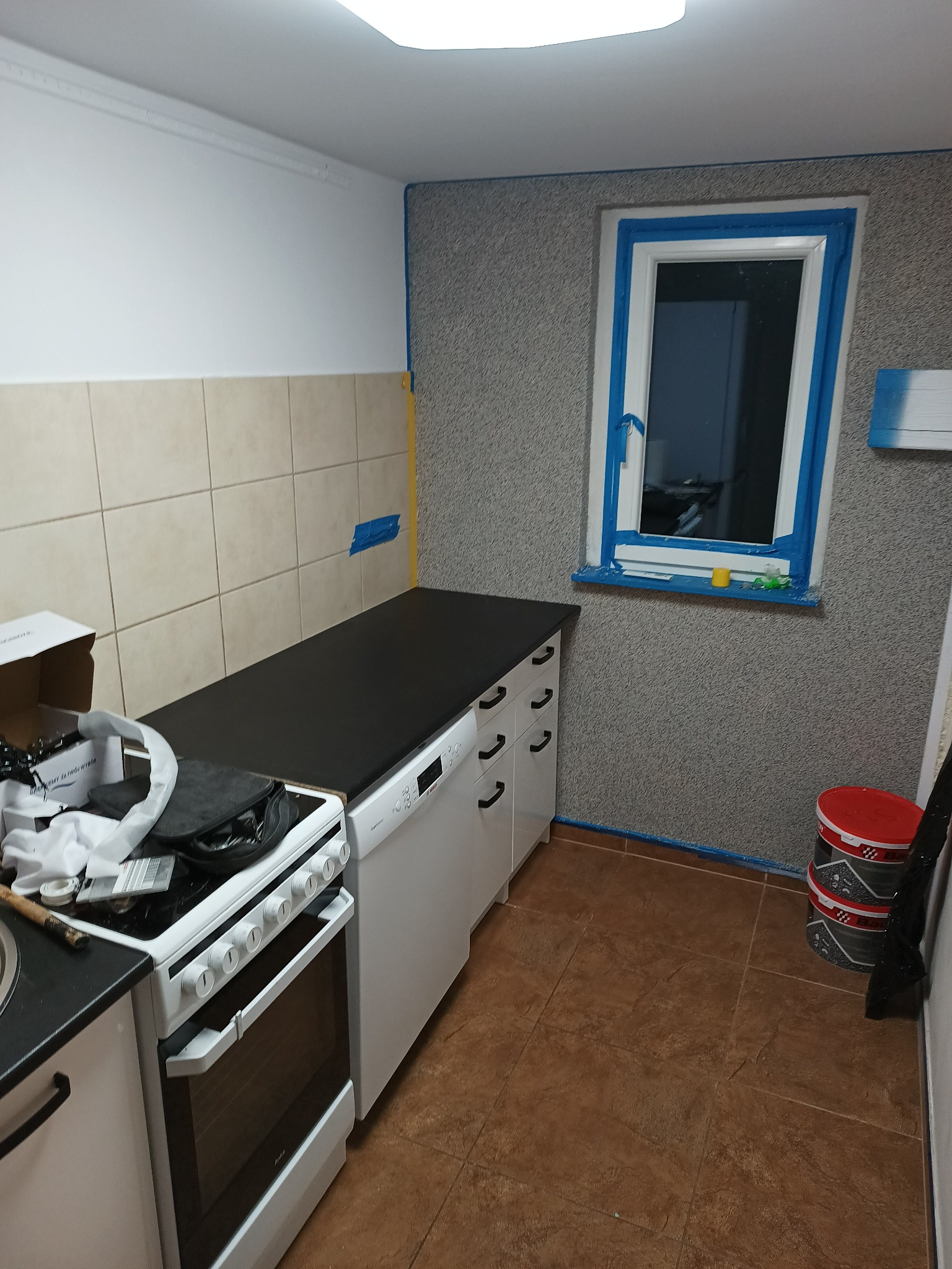 Kuchnia w trakcie odświeżania: ściany z fakturą zabezpieczone taśmą malarską wokół okna i przy podłodze, widoczne puszki z farbą, częściowo zakryte sprzęty AGD i karton z akcesoriami na kuchence.