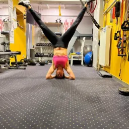 Kobieta w czarnych legginsach i różowym topie wykonuje pozycję stojąc na głowie z nogami rozwartymi w literę V, trzymając je na taśmach TRX w sali fitness z żółtymi ścianami i sprzętem do ćwiczeń.