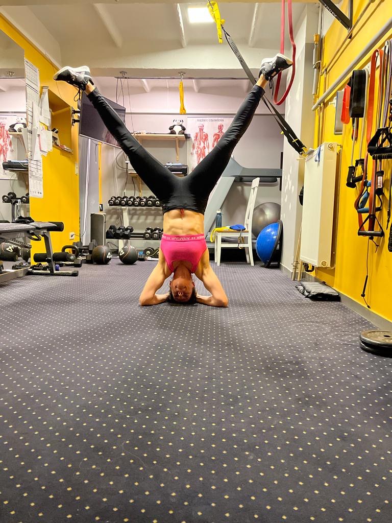 Kobieta w czarnych legginsach i różowym topie wykonuje pozycję stojąc na głowie z nogami rozwartymi w literę V, trzymając je na taśmach TRX w sali fitness z żółtymi ścianami i sprzętem do ćwiczeń.