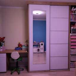 Mebl-Art Meble na Wymiar - Biała szafa przesuwna z lustrem w pokoju dziecięcym, obok biurko z zielonym krzesłem. W lustrze odbija się zabawkowy aneks kuchenny. Na półkach zabawki i książki.
