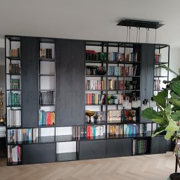 Mebl-Art Meble na Wymiar - Czarna, nowoczesna biblioteka na wymiar z otwartymi półkami i przesuwnymi drzwiami, wypełniona książkami i dekoracjami, oświetlona wiszącymi lampami.