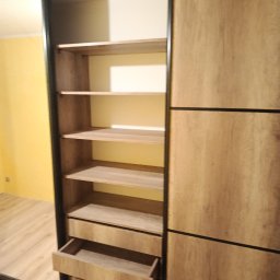 Mebl-Art Meble na Wymiar - Otwarta szafa wnękowa z przesuwnymi drzwiami, widoczne półki i wysunięta szuflada. Fronty w odcieniach drewna, czarne obramowanie. Żółta ściana w tle.