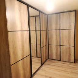 Mebl-Art Meble na Wymiar - Jasna szafa wnękowa z przesuwnymi drzwiami, wykończona drewnopodobnym dekorem i czarnymi ramami, z lustrem na jednym ze skrzydeł, w rogu pokoju.