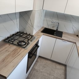 Mebl-Art Meble na Wymiar - Nowoczesna kuchnia z białymi szafkami i blatem imitującym drewno. Widoczna płyta gazowa, zlewozmywak i marmurowe wykończenie ściany. Ujęcie z góry, podkreślające ergonomię.