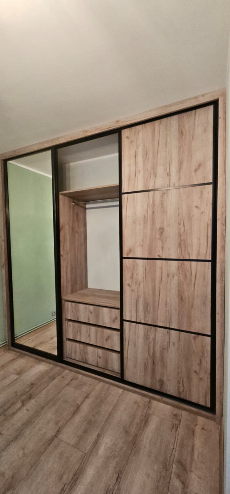 Szafa wnękowa z przesuwnymi drzwiami, wykończona okleiną drewnopodobną i czarnymi ramami. Widoczne półki, szuflady i lustro na drzwiach.