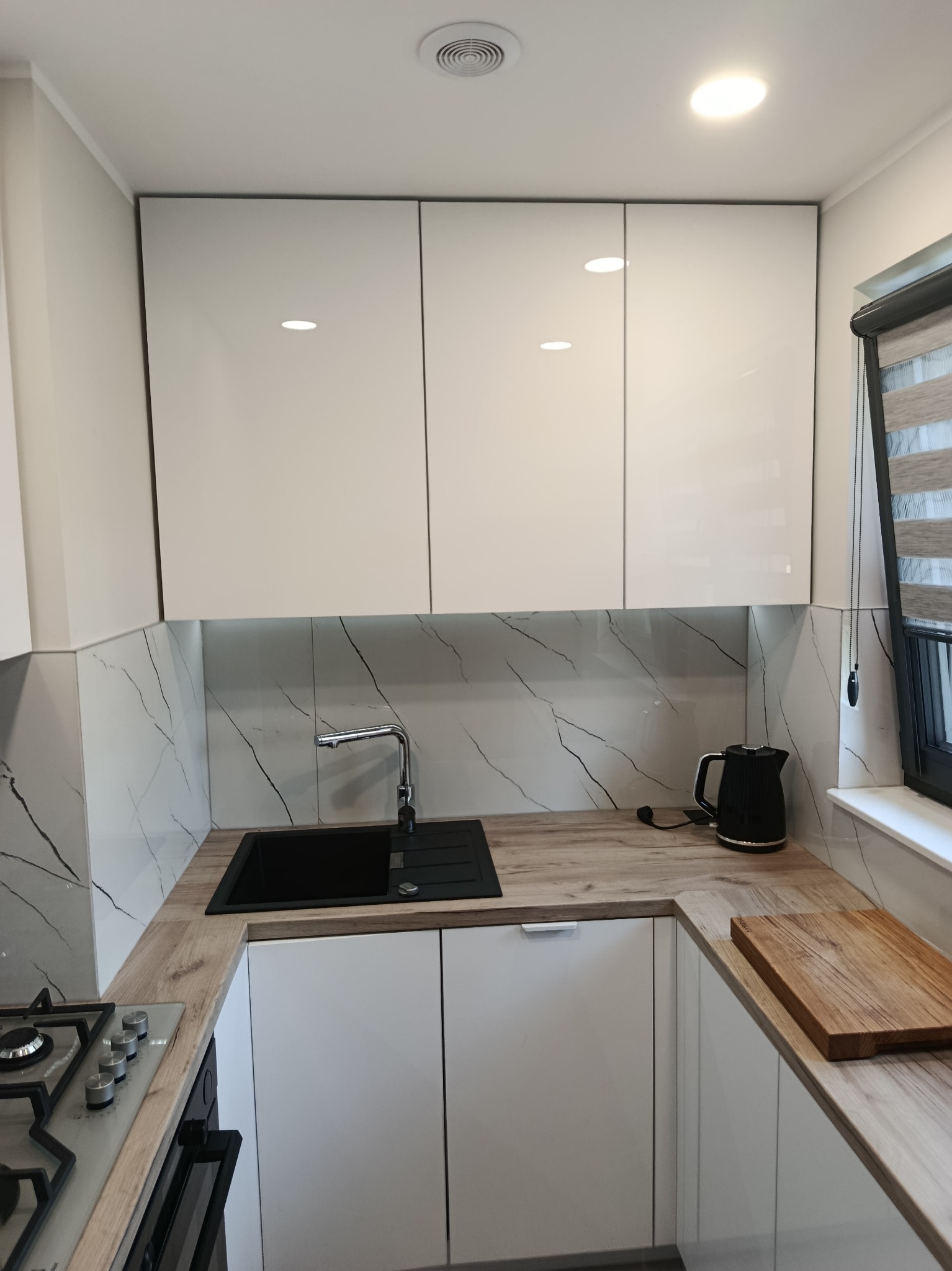 Nowoczesna kuchnia z białymi, lakierowanymi frontami, drewnianym blatem i marmurowymi płytkami na ścianie. Widoczny czarny zlew i płyta gazowa.