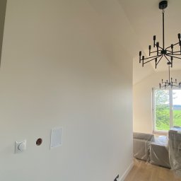 Stanislaw Myroshnychenko - Jasne, świeżo pomalowane wnętrze z odsłoniętymi gniazdkami i puszkami elektrycznymi na ścianie, zabezpieczonymi meblami folią ochronną i nowoczesnym czarnym żyrandolem.