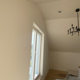 Stanislaw Myroshnychenko - Pomalowane na jasny odcień ściany i sufit pod skosem w pokoju z oknem i czarnym, nowoczesnym żyrandolem. Podłoga drewniana, zabezpieczona folią malarską.