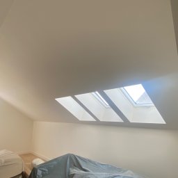 Stanislaw Myroshnychenko - Poddasze z trzema oknami dachowymi, ściany świeżo pomalowane na jasny kolor, podłoga częściowo widoczna, meble zabezpieczone folią malarską, karton na podłodze.