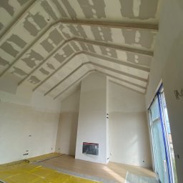 Stanislaw Myroshnychenko - Wnętrze domu w trakcie remontu: sufit z drewnianymi belkami i fragmentami gładzi, ściany po szpachlowaniu, podłoga zabezpieczona folią, widoczny kominek.