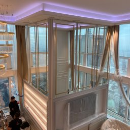 Stanislaw Myroshnychenko - Montaż szklanej konstrukcji z pozłacanymi ramami w nowoczesnym apartamencie z widokiem na miasto, pracownicy w czarnych uniformach na podnośniku, elementy wnętrza zabezpieczone folią ochronną.