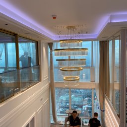 Stanislaw Myroshnychenko - Montaż wielopoziomowego, okrągłego żyrandola z kryształkami w luksusowym apartamencie z dużymi oknami i widokiem na miasto, dwóch monterów na drabinie.