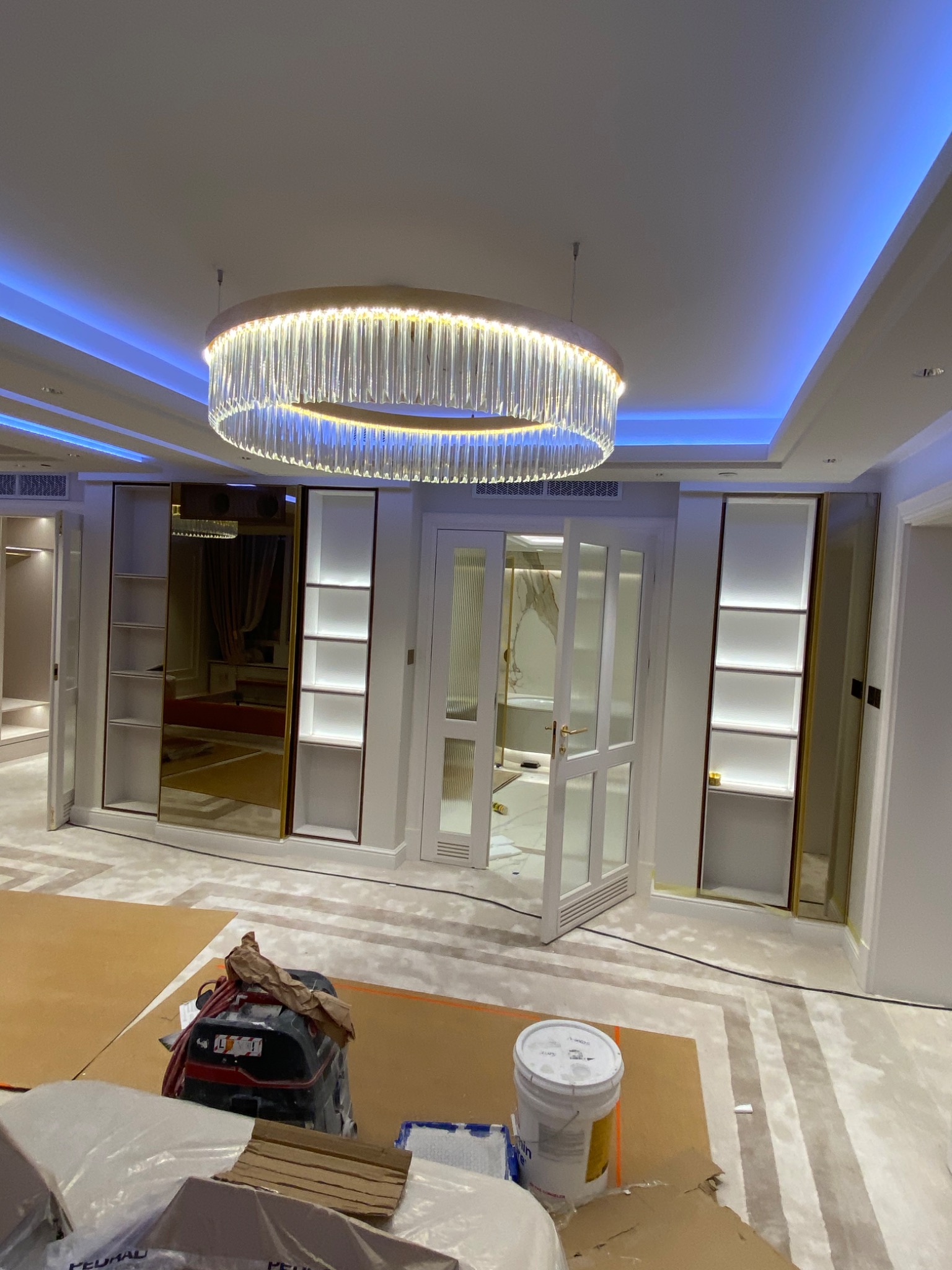 Przygotowanie do prac wykończeniowych w apartamencie w Warszawie: zabezpieczona podłoga, wiadro z farbą i szlifierka w luksusowym wnętrzu z designerskim oświetleniem i wbudowanymi szafami.