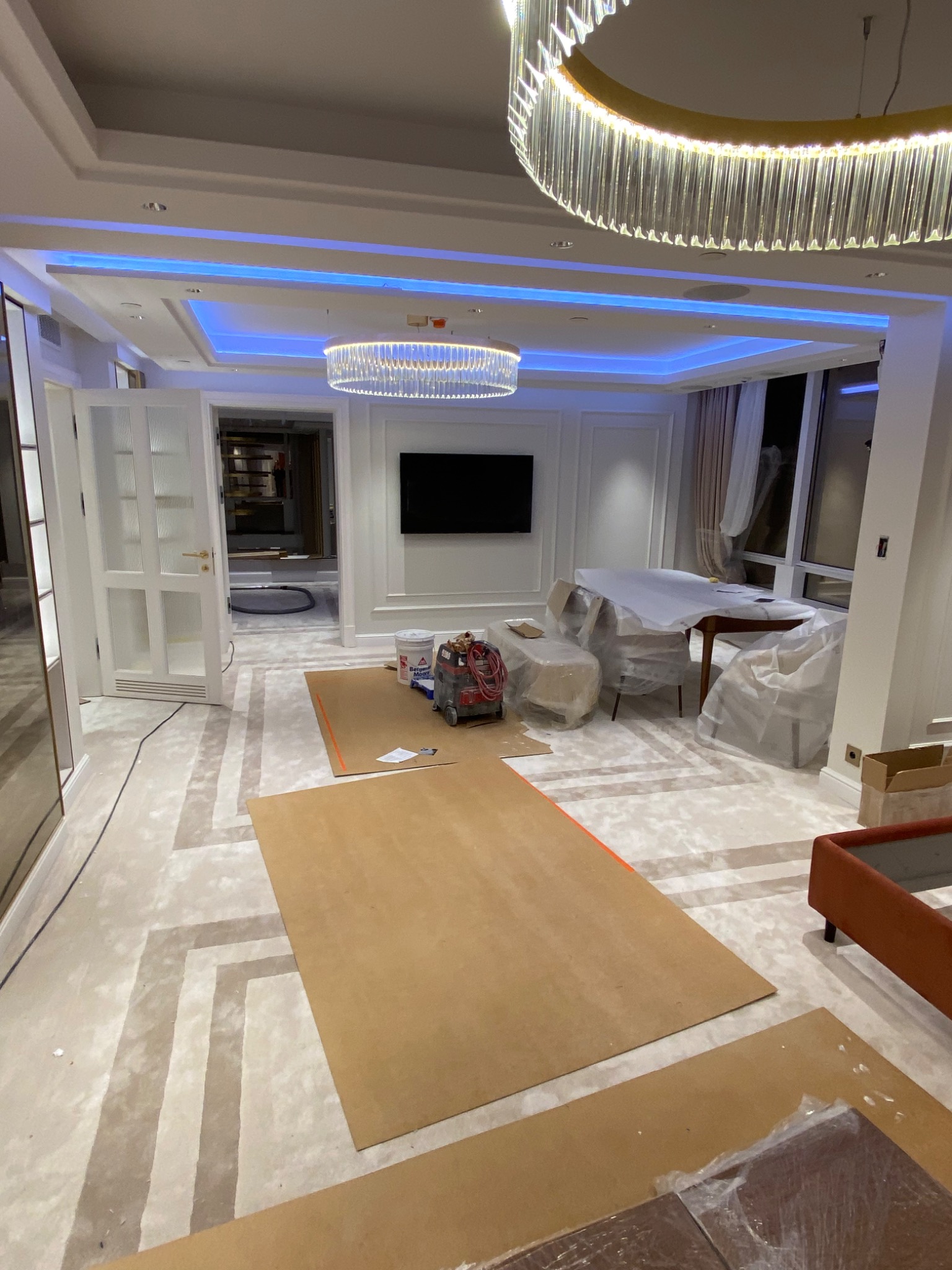 Luksusowe wnętrze w trakcie zabezpieczania podłogi kartonem przed pracami wykończeniowymi, widoczny sprzęt do prac na ścianach, meble zabezpieczone folią, nowoczesne oświetlenie sufitowe.