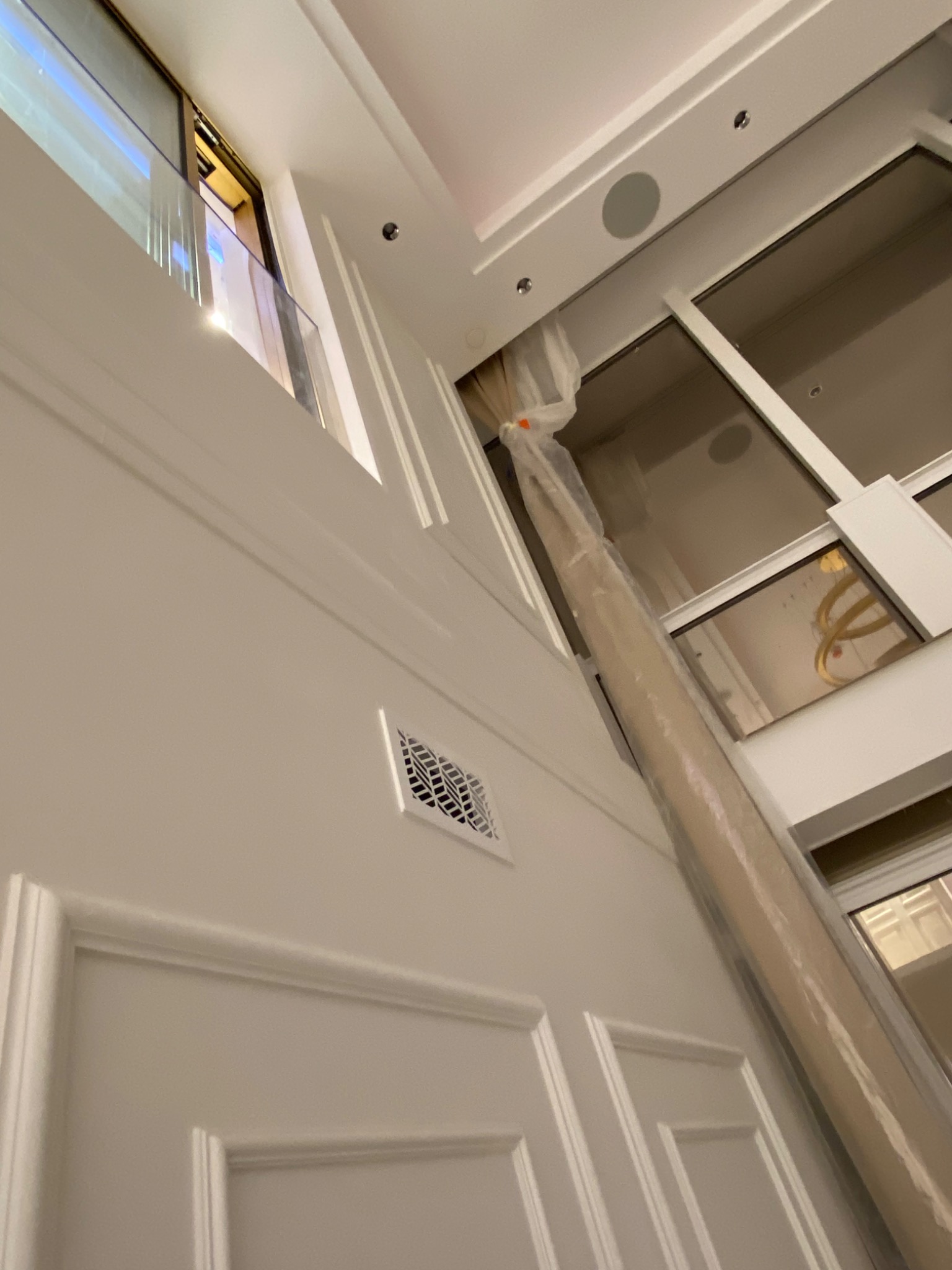 Wnętrze w trakcie remontu: ściana z dekoracyjnymi panelami, kratka wentylacyjna i okno z widocznym zabezpieczonym folią elementem, prawdopodobnie kolumną lub rurą.