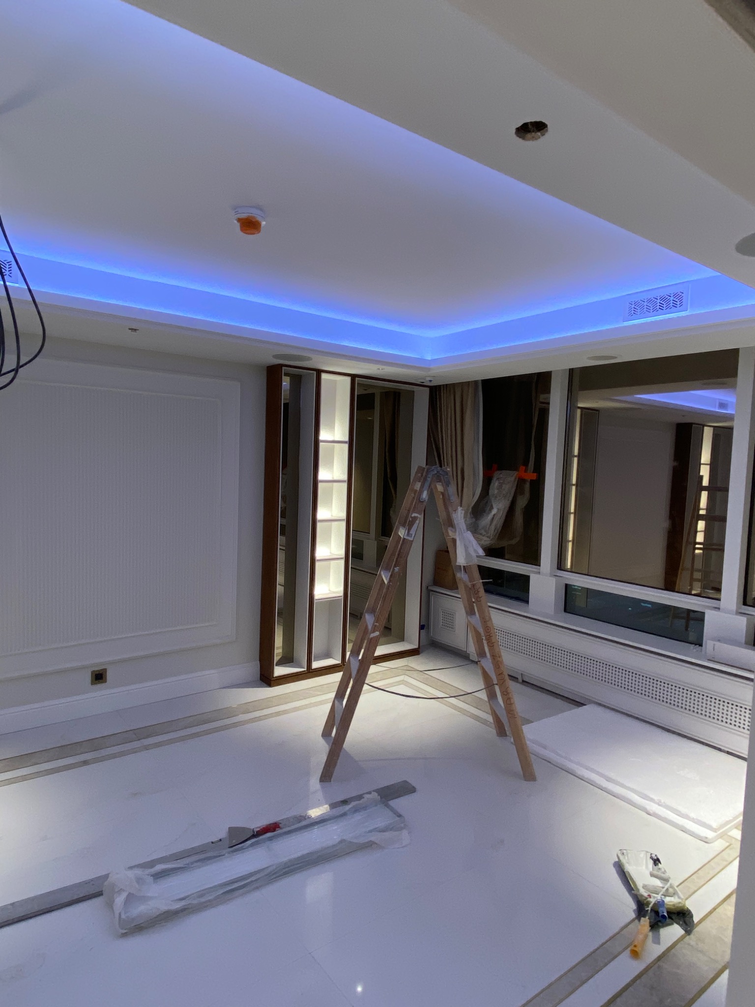 Wnętrze w trakcie prac wykończeniowych: drewniana drabina oparta o ścianę z dużym oknem, pod sufitem niebieskie oświetlenie LED, na podłodze rozłożone materiały budowlane i narzędzia malarskie.