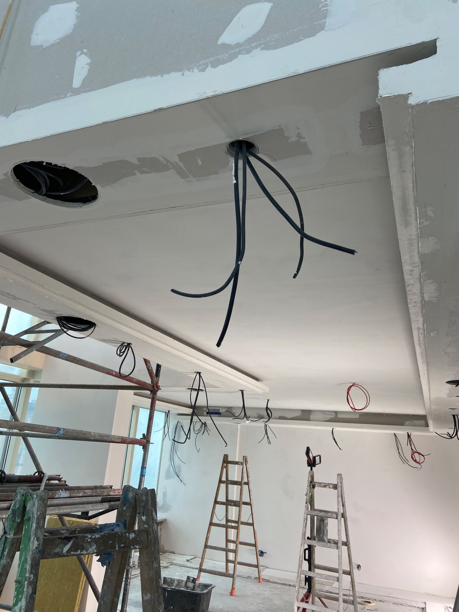 Sufit podwieszany w trakcie montażu z widocznymi kablami elektrycznymi, rusztowaniami i drabinkami w tle, ukazujący etap prac remontowych.