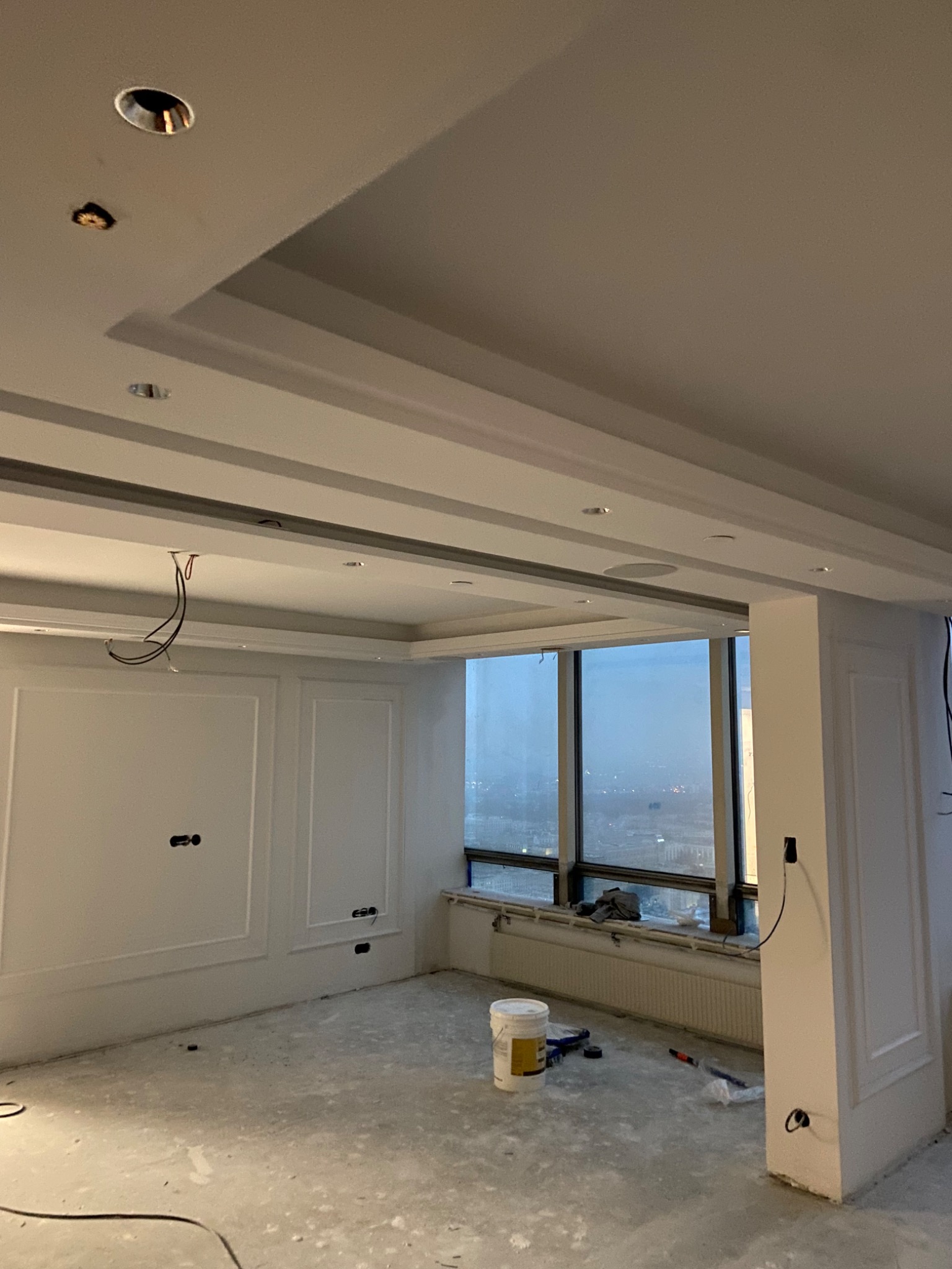 Wnętrze w trakcie prac wykończeniowych: surowe ściany z dekoracyjnymi panelami, odsłonięte kable, widok na miasto zza okna, wiadro farby i narzędzia na podłodze.