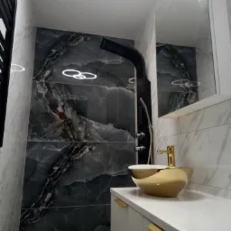 Nowoczesna łazienka z czarną armaturą prysznicową, złotą umywalką nablatową i marmurowymi płytkami w odcieniach szarości.