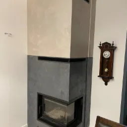 Nowoczesny kominek narożny z dwukolorową obudową (beż i szary), czarny wkład kominkowy, marmurowe płytki podłogowe, wiszący zegar z wahadłem na ścianie.