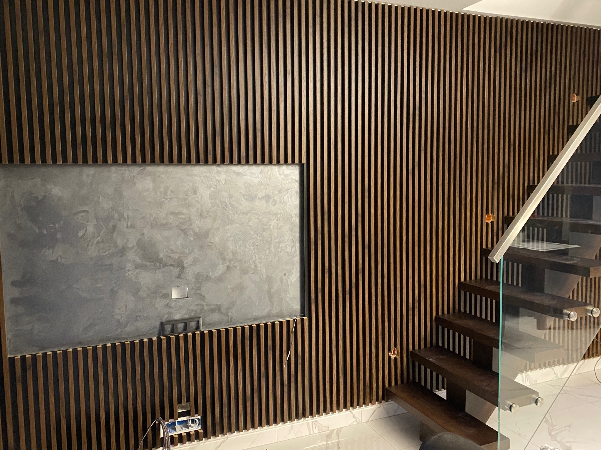 Drewniane lamele na ścianie z wnęką na telewizor z szarą ścianą dekoracyjną i drewniane schody ze szklaną balustradą.