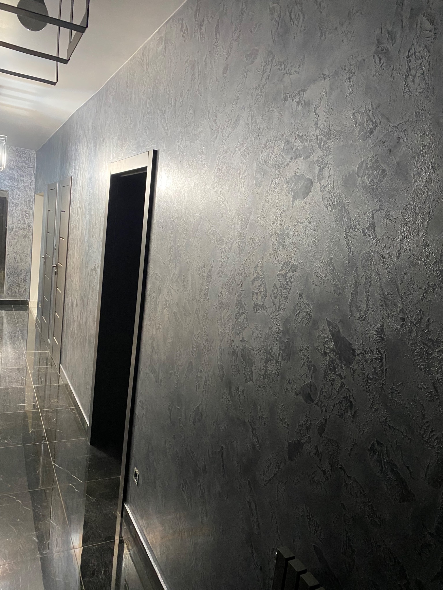 Korytarz z dekoracyjną, szarą ścianą o nieregularnej fakturze, ciemną podłogą z połyskiem i czarnymi futrynami drzwi.