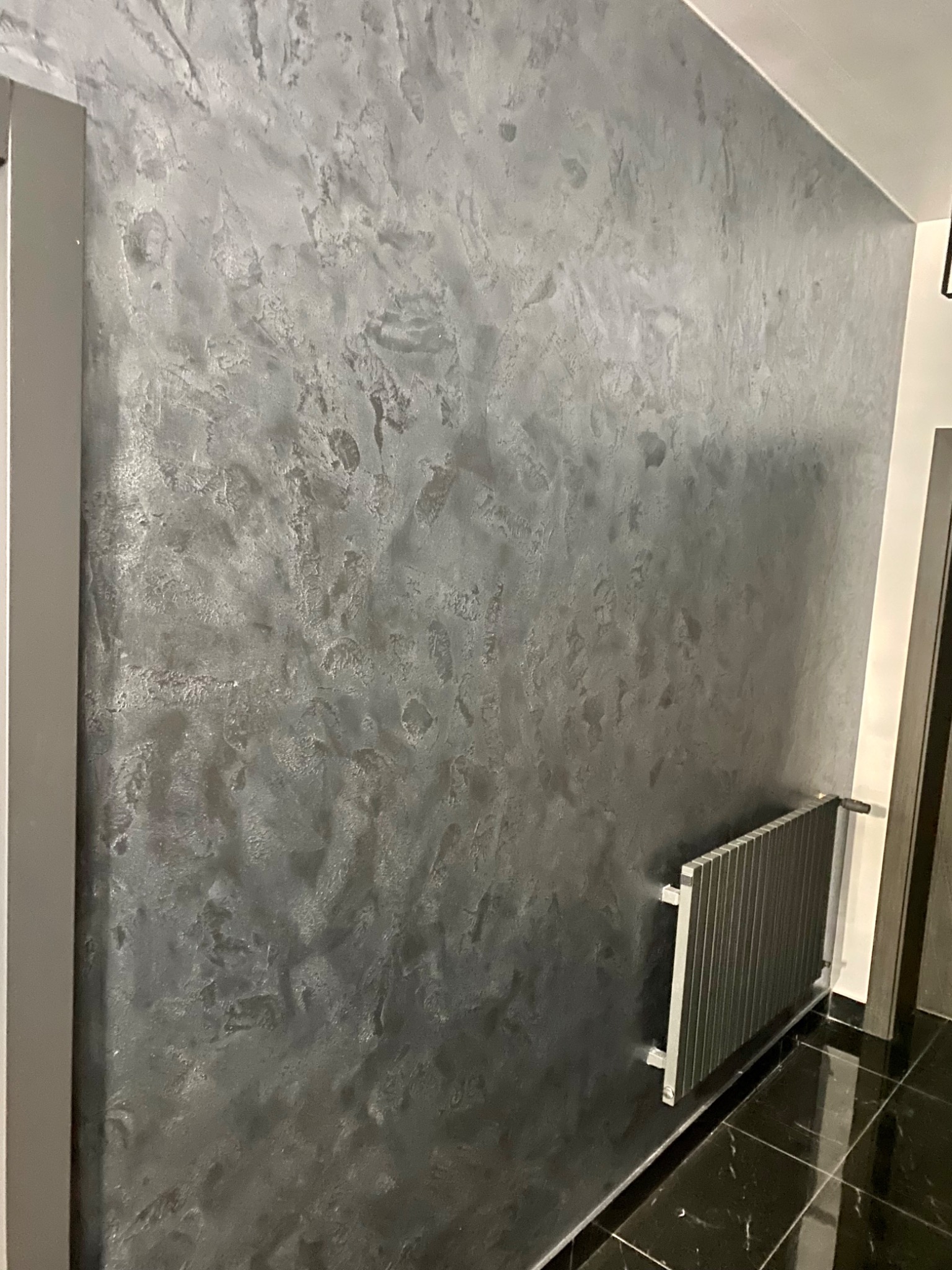 Ściana z dekoracyjną, ciemną szarą fakturą, widoczny grzejnik i fragment podłogi z czarnego marmuru.